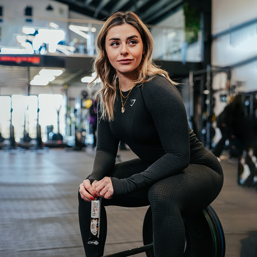 Eine Frau sitzt auf den Gewichten in einem Fitnessstudio und hält einen nährstoffreichen Süßwaren-Proteinriegel